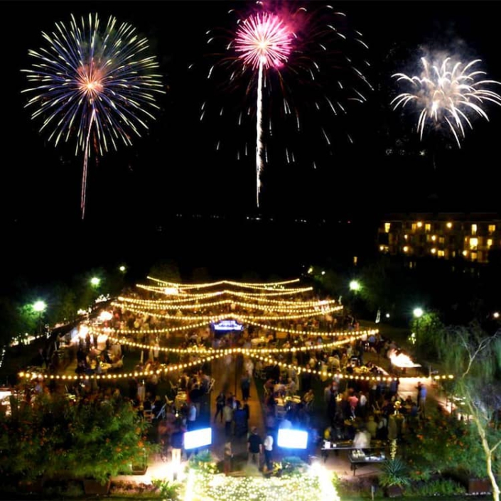 A nighttime outdoor celebration with string lights and a large crowd, as colorful fireworks light up the sky above.
